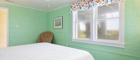 A room with a bed, a window with a butterfly-patterned valance, a ceiling fan, and a wicker chair.