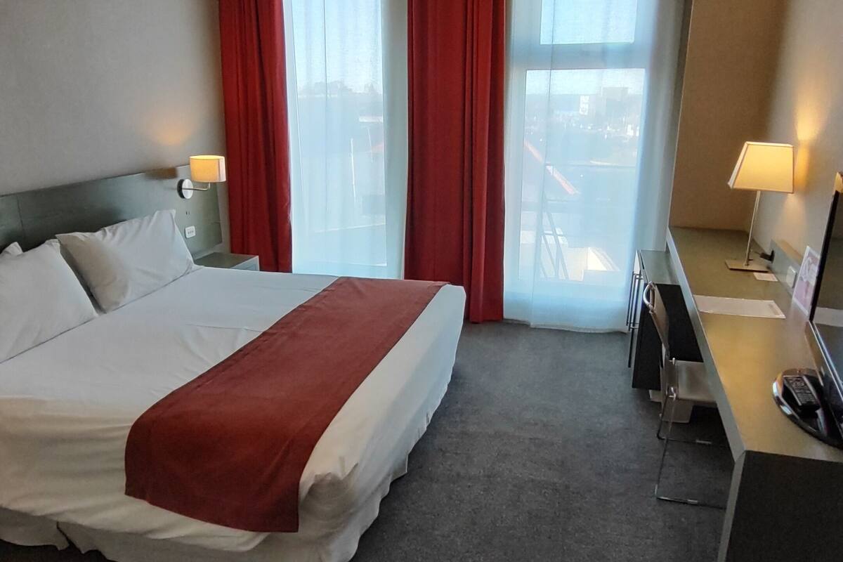 Una habitació d'hotel amb llit, escriptori, cadira, làmpada i finestra amb cortines.