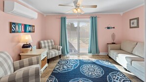 Una sala de estar acogedora con un ventilador de techo, un letrero 'Sail Away', un tapete estampado y vistas a través de la puerta de vidrio.