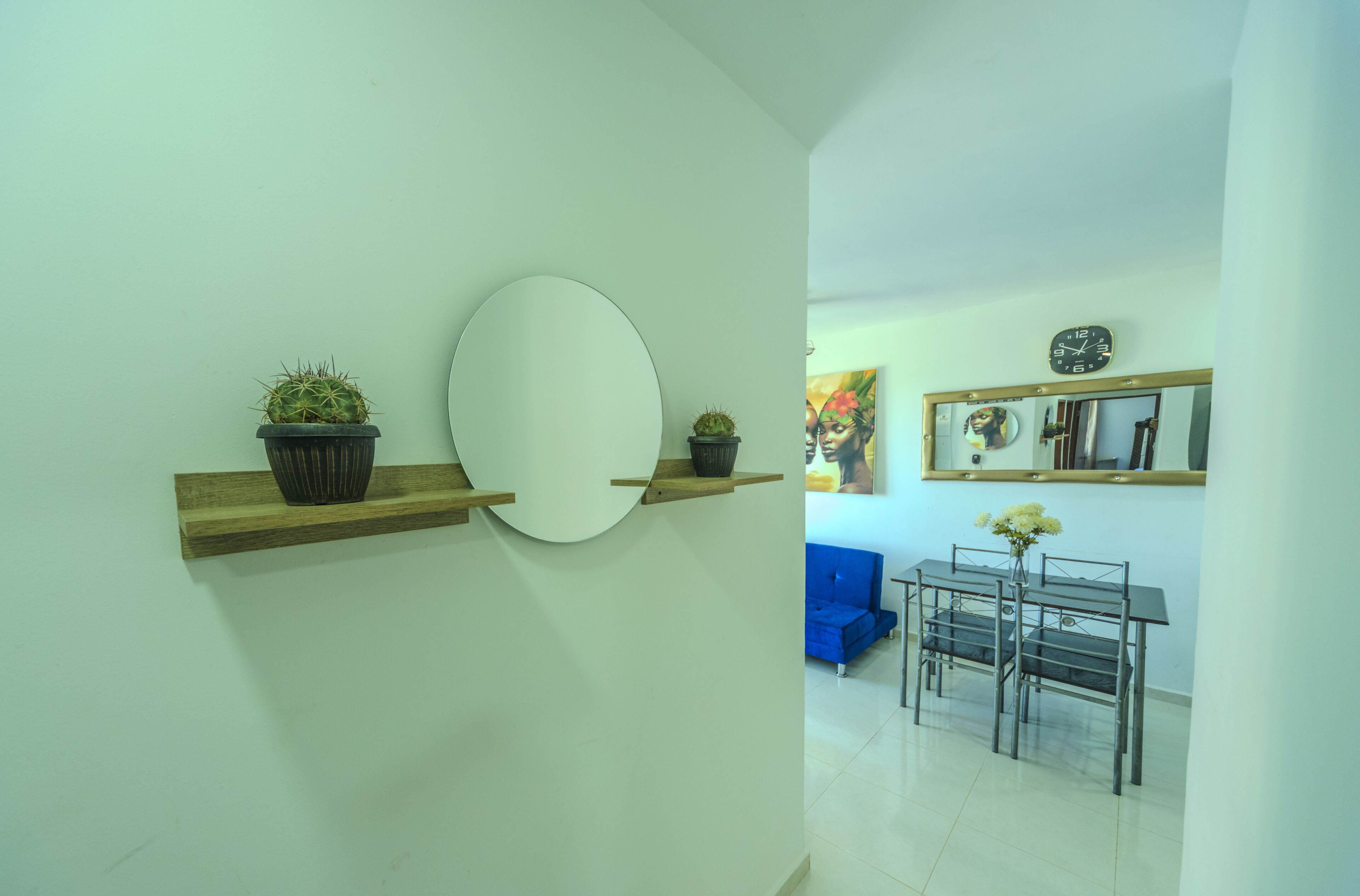 Un intérieur moderne avec un grand miroir rond, des étagères en bois ornées de plantes en pot, un coin repas avec une table et des chaises en verre, et un canapé bleu.