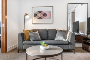 A living room with a grey sofa, a round coffee table with a plant, a floor lamp, and a framed abstract painting on the wall.