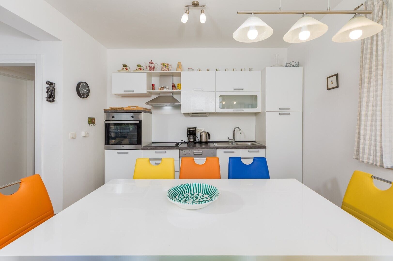 A modern kitchen with white cabinets, a microwave, an oven, and a coffee maker. There are colorful chairs and a white table with a green and white plate.