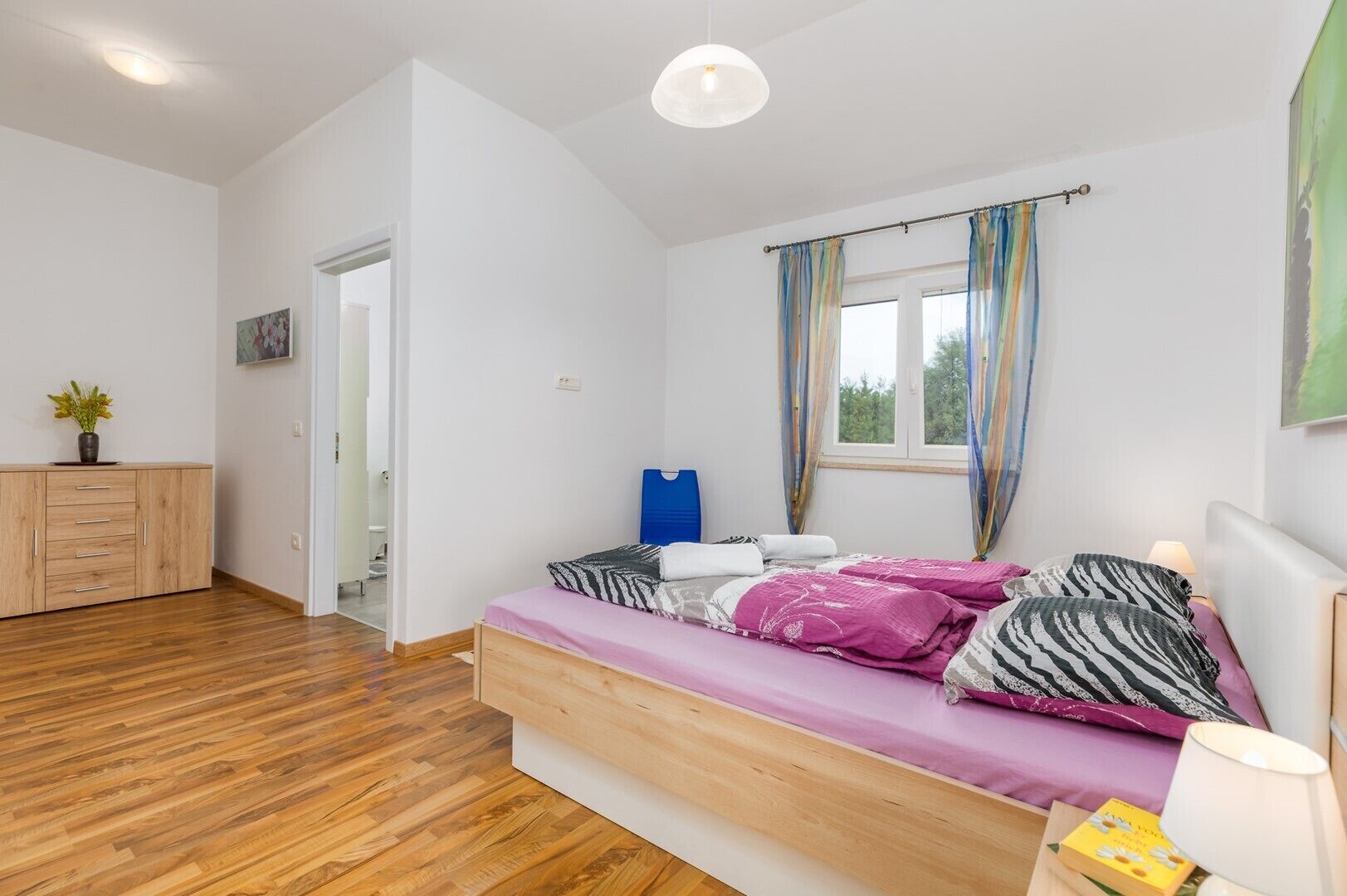 Camera da letto con un letto, un comodino, una lampada, una finestra con tende, una porta e un settimino in legno.