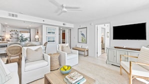 A spacious living room with white furniture, a flat-screen TV, and a dining area with a kitchen in the background.