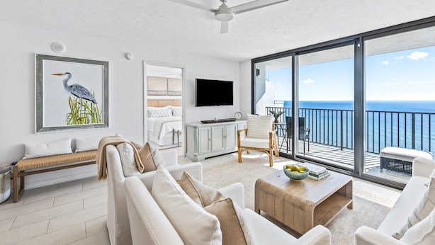 A modern living room with a large window, a ceiling fan, a flat-screen TV, and a balcony with a view of the ocean.