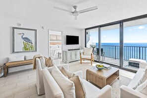 A modern living room with a large window, a ceiling fan, a flat-screen TV, and a balcony with a view of the ocean.