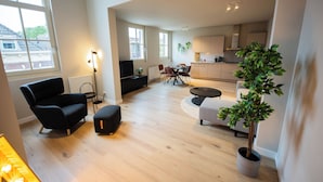 A spacious living room with a large window, a flat-screen TV, a dining area with a table and chairs, and a kitchen in the background.