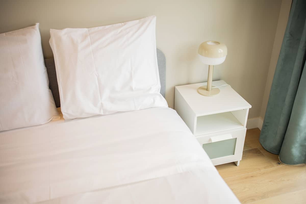 A neatly made bed with white bedding and pillows, a white bedside table with a lamp, and a grey curtain.