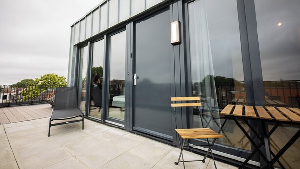 A modern building with large glass doors and windows, a balcony with a chair and a small table, and a view of the surrounding area.