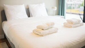 A neatly made bed with white linens and towels, a bedside table with a lamp, and a view of a house through a window.