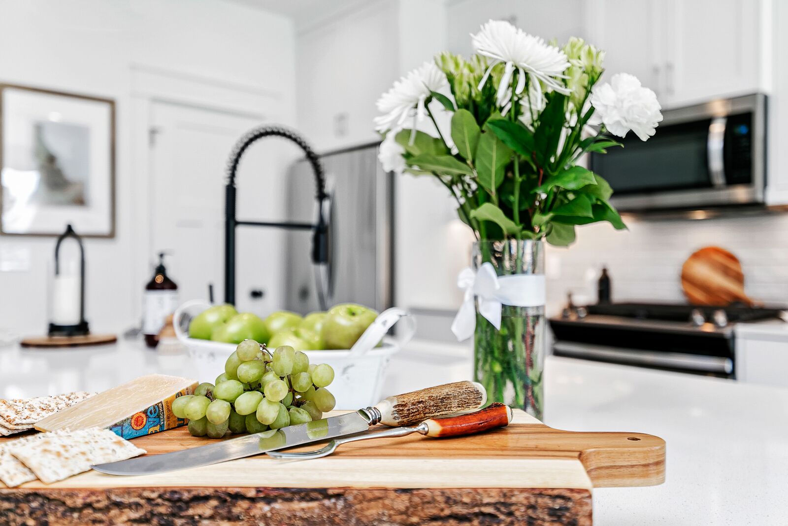 A kitchen counter with a knife, grapes, a cutting board, a bowl of apples, and a vase of flowers.