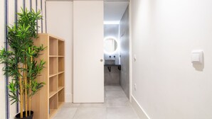 A hallway with a bamboo plant, a wooden shelf unit, and a bathroom with a round mirror and modern fixtures. - Bom Retiro's Duplex by LovelyStay (Porto)