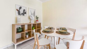Un comedor con una mesa redonda dispuesta para cuatro personas, rodeada de sillas blancas. Hay una estantería de madera con varios objetos decorativos y un pequeño ventilador sobre la mesa.