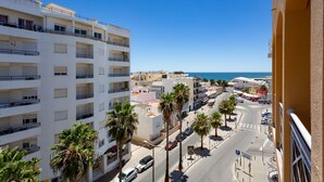 Un hotel de varios pisos con balcones, una calle con palmeras y vista al mar.