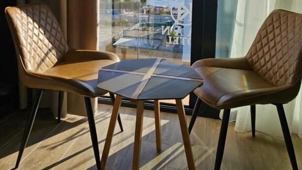 A modern room with two leather chairs and a small table, large windows with a view of a body of water and buildings, and a patterned carpet. - KAAN Hotel (Orikum)