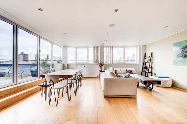 A spacious living area with a dining table and chairs, a sofa, and a cart. Large windows provide a view of the cityscape.