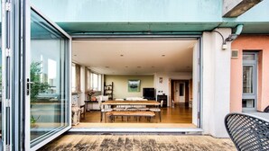 An open-plan living area with a dining table and benches, a kitchen area with a stove and cabinets, and a sliding glass door leading to an outdoor patio. - GuestReady – Stylish 2BR in Angel with Balcony (London)