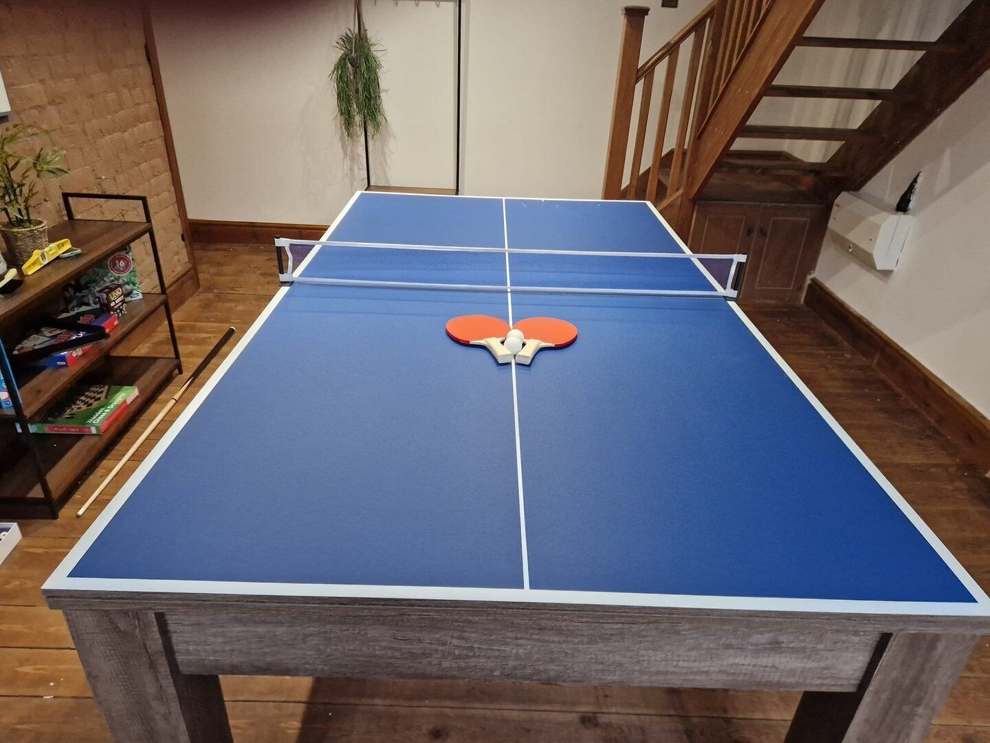 A ping pong table with two paddles in the center, a wooden floor, and a staircase in the background.