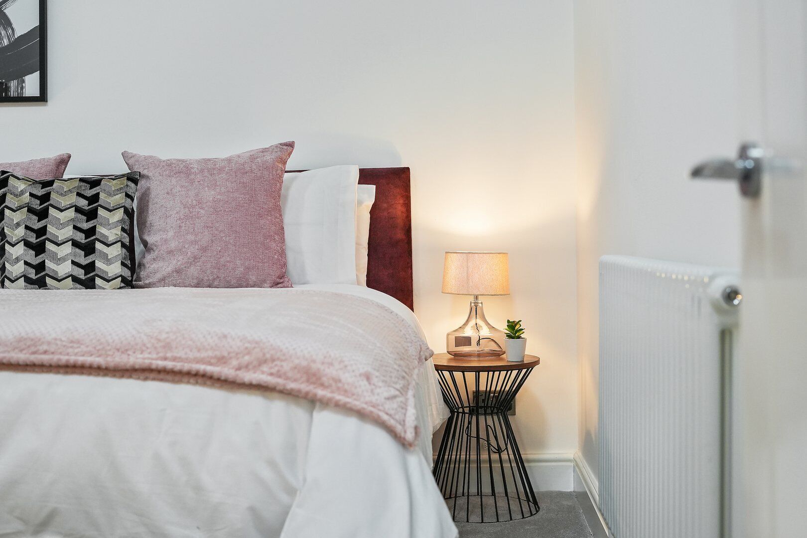 A neatly made bed with a white comforter and pink pillows, a bedside table with a lamp and a small plant, and a radiator on the wall.