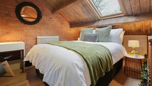 A bedroom with a wooden ceiling, a brick wall, a skylight, a bed with white bedding, green pillows, a bedside lamp, and a radiator.