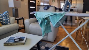 Una sala de estar con un sofá, una mesa de centro con libros, una tabla de planchar y una cocina al fondo.
