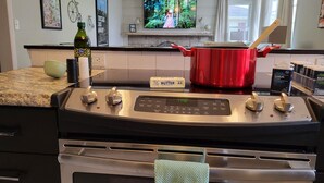 Una cocina con estufa de acero inoxidable, una olla roja, una botella de aceite de oliva y un televisor sobre la estufa.