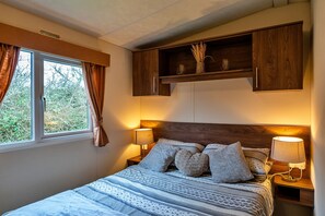 Un dormitorio con cama, dos lámparas de noche, ventana con cortinas, estante de madera sobre la cama y vistas a zonas verdes.