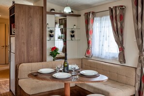 Un comedor con una mesa redonda preparada para cuatro personas, un sofá y una pequeña cocina con un refrigerador y estanterías.