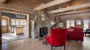 Ein Blockhaus-Interieur mit Kamin aus Stein, Holzböden, einem roten Samtsofa und einer Küchenzeile mit Holzausbau und einem großen Fenster.