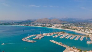 An aerial view of a coastal marina with boats docked, a marina building, and a beach area with buildings in the background. - House Massanet for 8 with swimming pool near the beach and all amenities (Puerto de Alcudia)