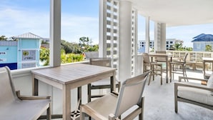 Une terrasse extérieure avec des tables et des chaises en bois, de grandes fenêtres et une vue sur les bâtiments et la verdure.