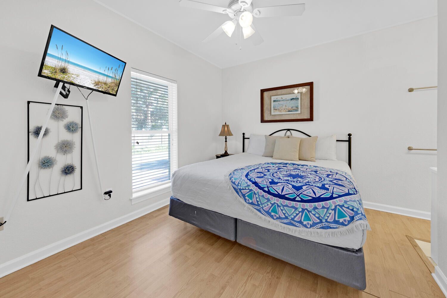 A bedroom with a bed, a TV mounted on the wall, a ceiling fan, a window with blinds, and a framed picture on the wall.