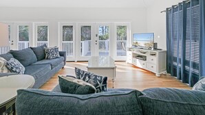 Amplia sala de estar con un sofá gris, una mesa de centro y un televisor sobre un mueble blanco. Amplias puertas de cristal dan a un balcón con vistas a árboles.