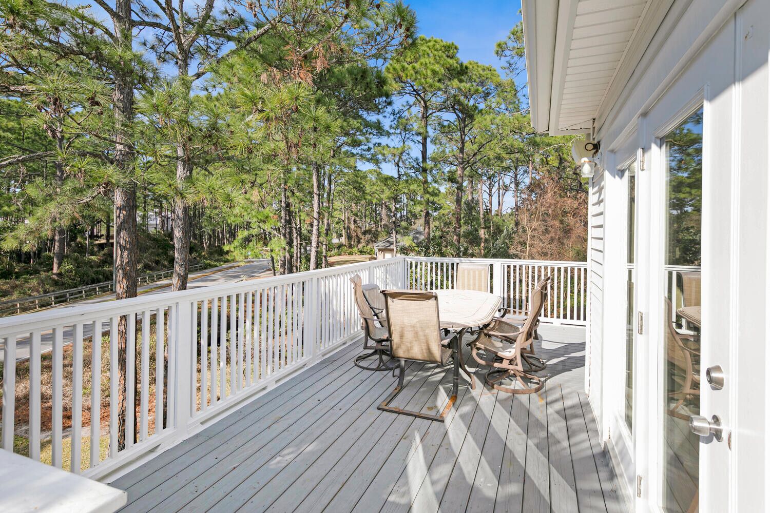 A wooden deck with a table and chairs, surrounded by a white railing, overlooking a forested area.
