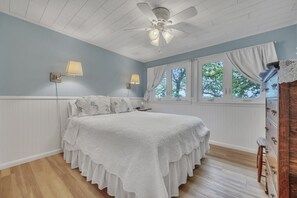 Ein Schlafzimmer mit einem Bett, zwei Wandlampen, einem Deckenventilator, einem Fenster mit Vorhängen und einer Kommode mit Schubladen.