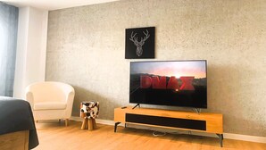 Ein modernes Wohnzimmer mit einem Flachbildfernseher auf einem Holzständer, einem weißen Sessel und einem kleinen Hocker aus Kuhfell.