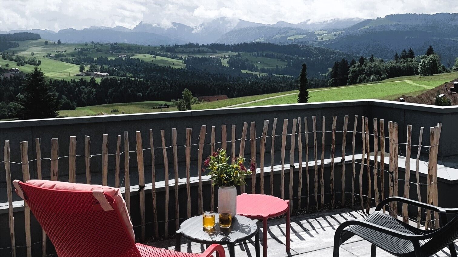 A balcony with a view of a mountainous landscape, a red chair, a black chair, and a small round table with a vase of flowers and two glasses.