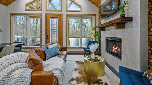 A cozy living room with a fireplace, leather sofas, a large window, and a TV mounted on the wall. - Private Pond | Kayaks · Spa Bath | Romantic Cabin (Broken Bow)