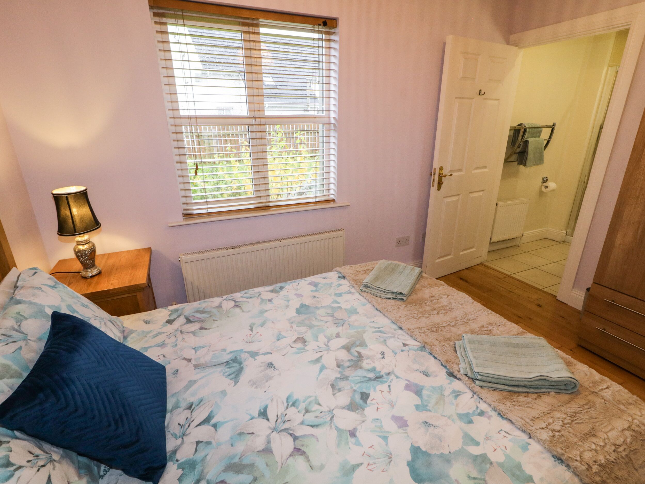 A bedroom with a bed, a nightstand, a window with blinds, a radiator, and a door leading to a bathroom.