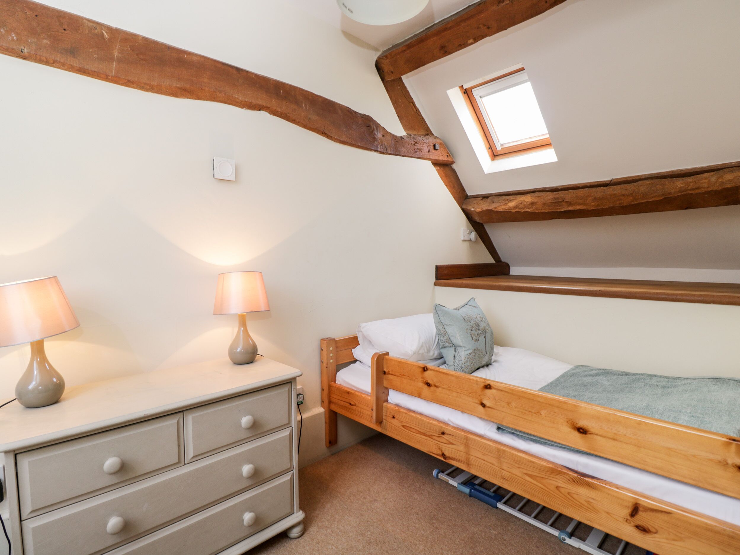 A bedroom with a wooden bunk bed, a bedside table with lamps, a dresser, and a skylight.