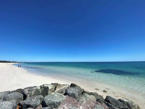 Une plage à l’eau claire, avec un rivage sablonneux et de gros rochers au premier plan.