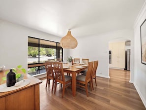 A dining area with a wooden table and chairs, a hanging light fixture, and a view through a large window.