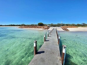 A pier extending into clear water with a sandy beach and trees in the background.