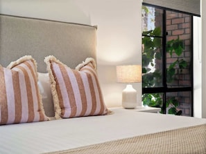 Une chambre d’hôtel avec un lit, des oreillers à rayures, une lampe de chevet et une vue sur la verdure par la fenêtre.