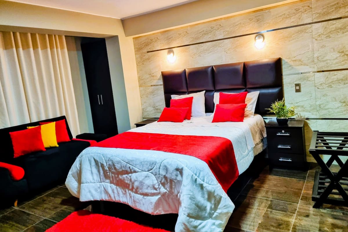 A modern bedroom with a large bed, a black headboard, red and white bedding, a black sofa with red cushions, a small table with a plant, and a wardrobe.