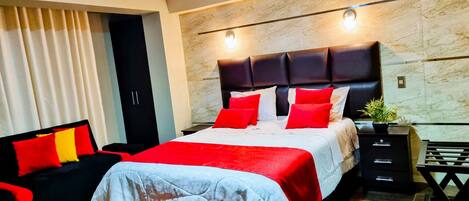 A modern bedroom with a large bed, a black headboard, red and white bedding, a black sofa with red cushions, a small table with a plant, and a wardrobe.