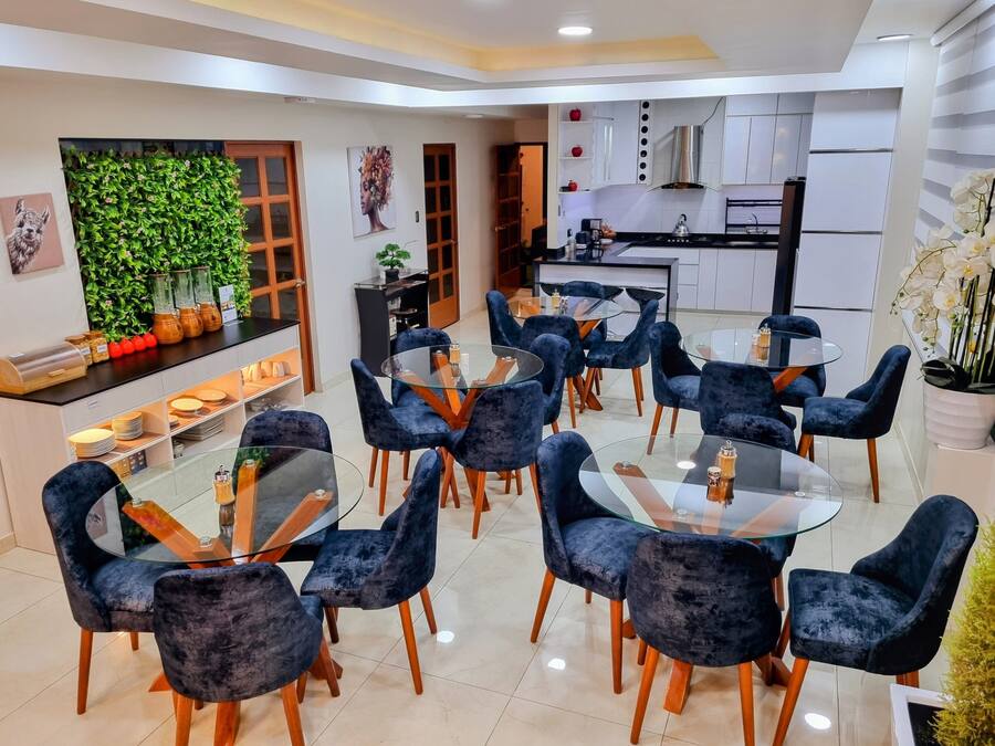 Un comedor moderno con mesas redondas de vidrio y sillas de terciopelo azul oscuro, una cocina al fondo y una pared verde con plantas.