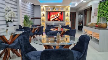Un soggiorno moderno con un tavolo da pranzo in vetro circondato da poltrone in velluto blu scuro, un camino con una TV e una mensola decorativa con diversi oggetti.