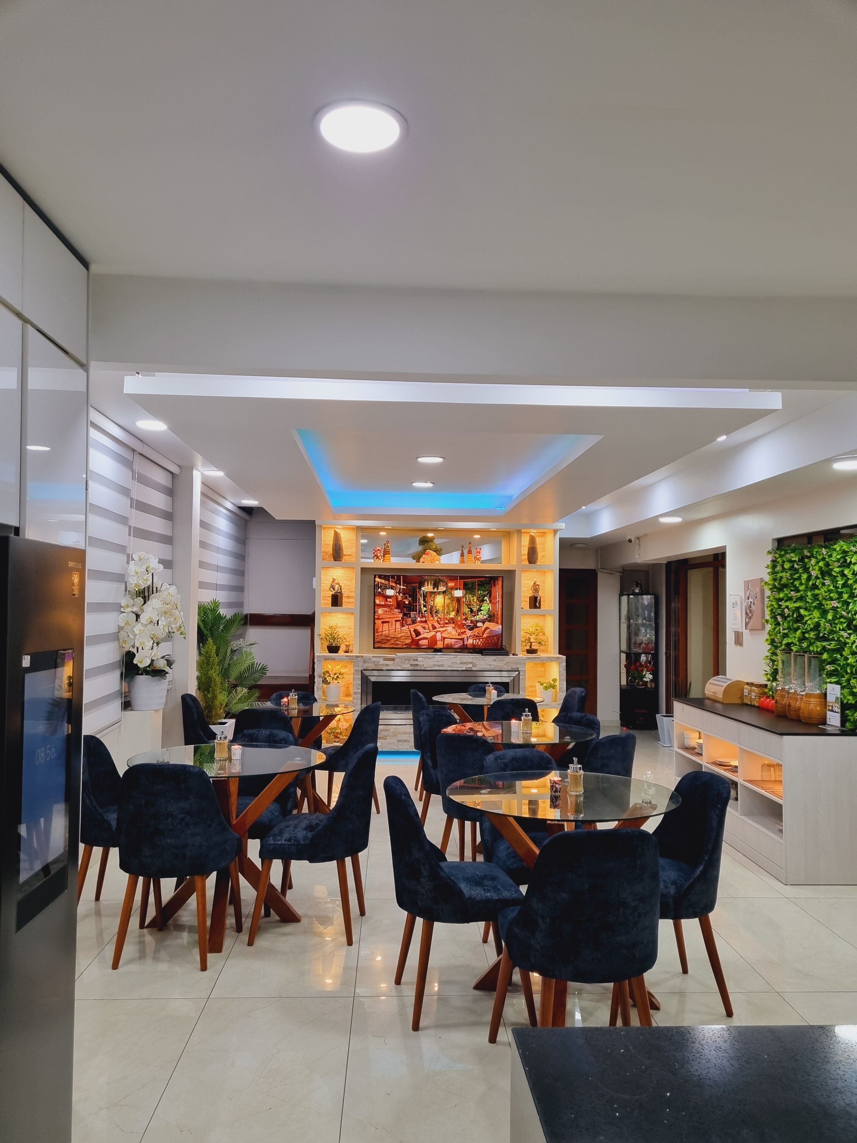Un comedor moderno con mesas redondas y sillas azul oscuro, una chimenea central y una pantalla de televisión mostrando una escena de restaurante.