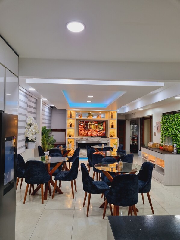 A modern dining area with round tables and dark blue chairs, a central fireplace, and a TV screen displaying a restaurant scene. - Hotel del Valle (El Tambo)